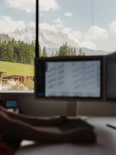 Eine silhouettierte Person sitzt an einem Schreibtisch mit zwei Monitoren; durch das Fenster Blick auf Berglandschaft mit Wald.