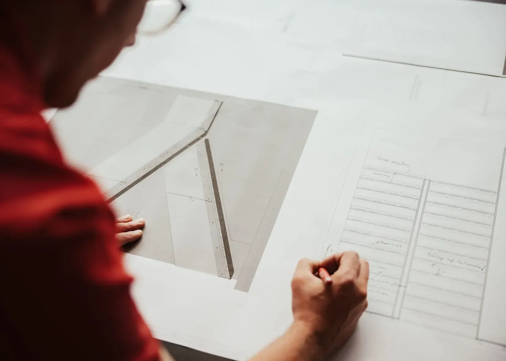 Person in roter Arbeitskleidung bearbeitet große technische Zeichnungen eines Metallbauunternehmens aus Südtirol; eine Hand liegt auf dem Blatt, die andere zeichnet.