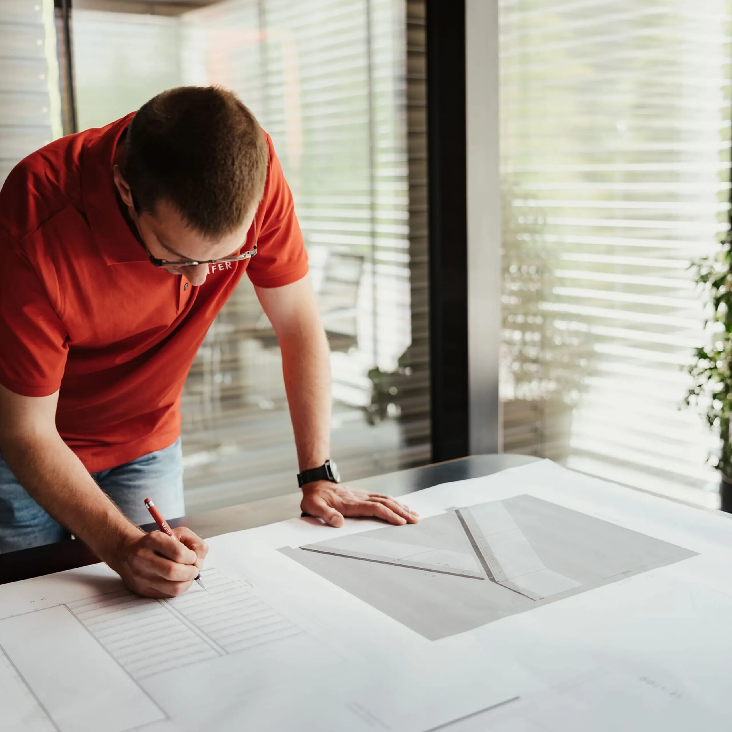 Eine Person in rotem Shirt beugt sich über große Baupläne und skizziert mit Bleistift; Glasfenster, Schreibtisch und Pflanze im Hintergrund.