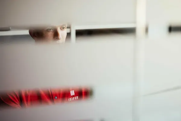 Person im roten Arbeitshemd mit ISIFER-Logo blickt durch unscharfe horizontale Lamellen; metallische Werkstattumgebung im Hintergrund.