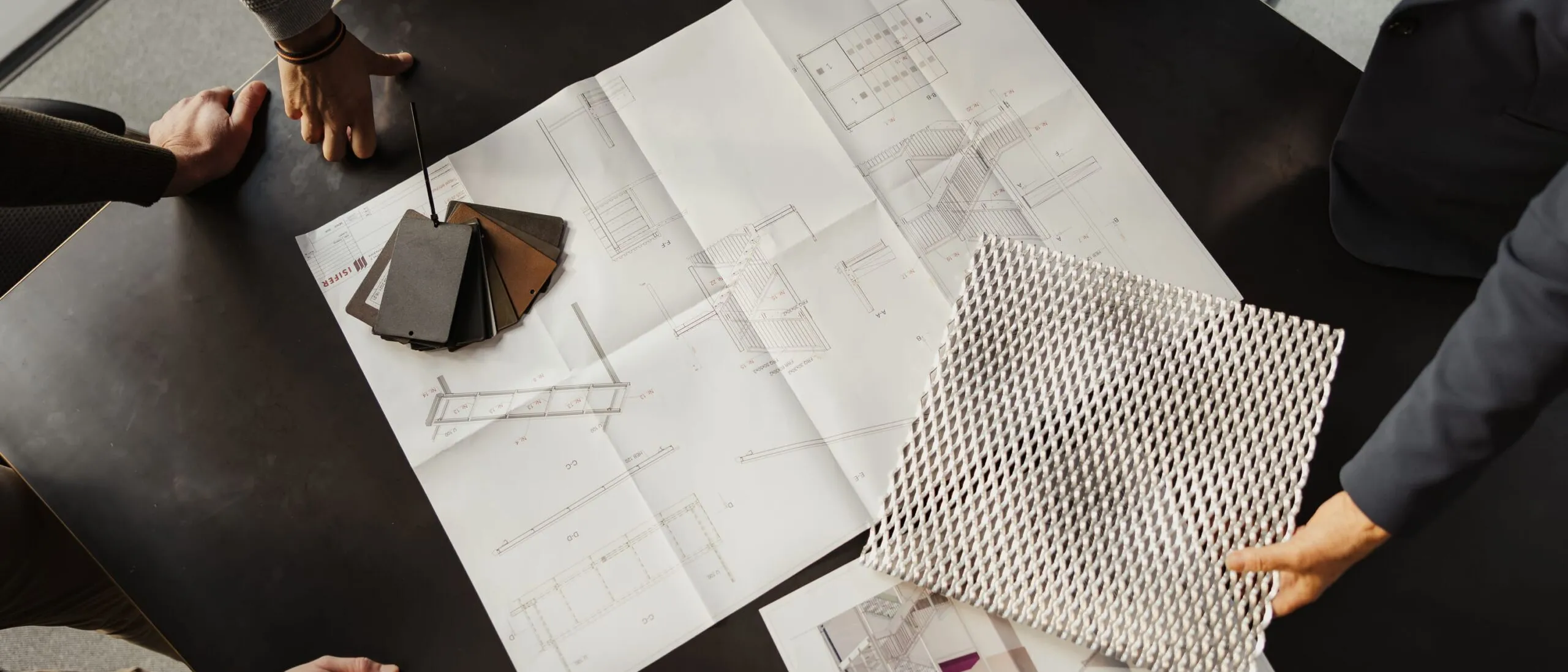 Gruppe von Personen bespricht Baupläne auf einem schwarzen Tisch; Farbmuster, Musterplatten und eine Metallgitter-Probe liegen sichtbar.