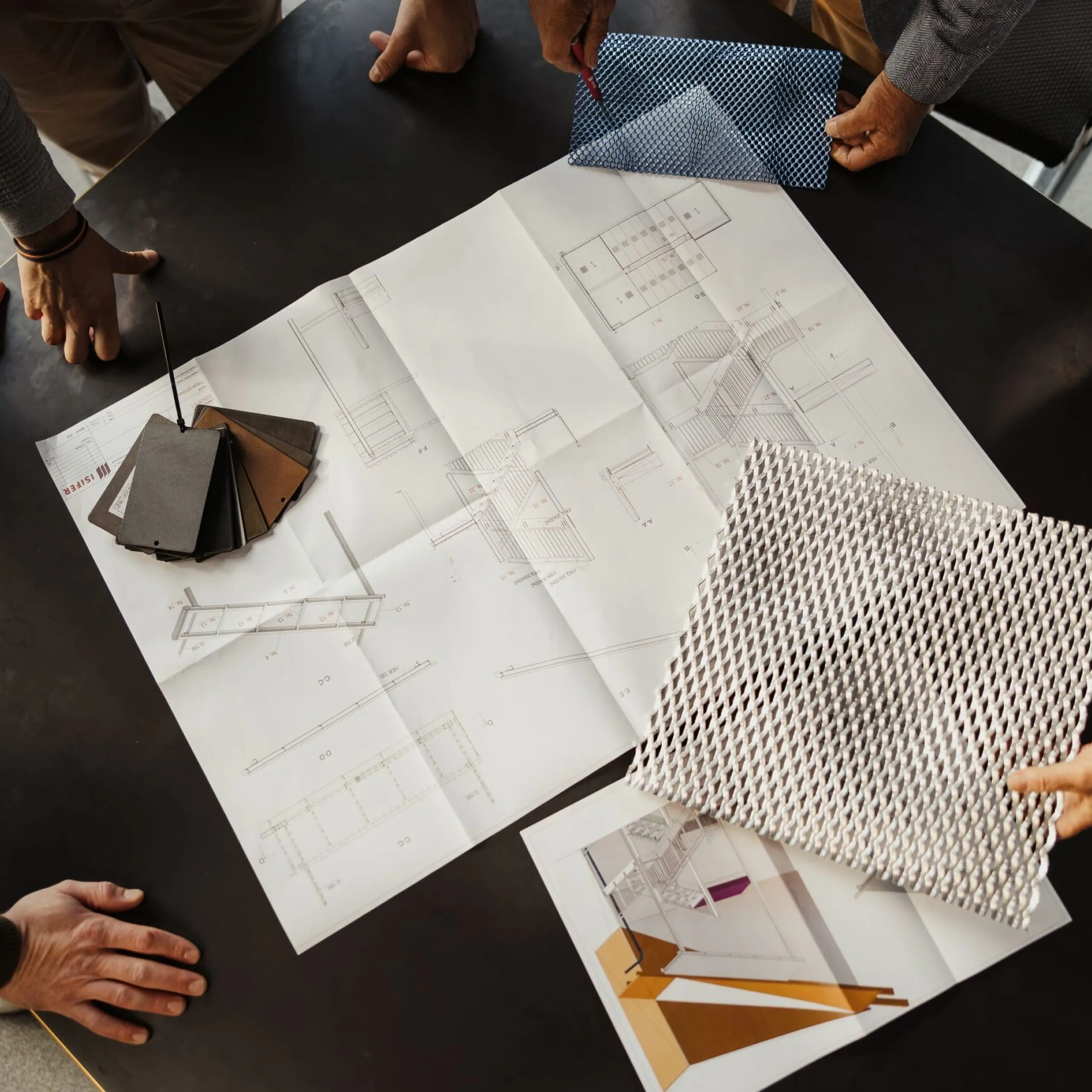 Gruppe von Personen bespricht Baupläne auf einem schwarzen Tisch; Farbmuster, Musterplatten und eine Metallgitter-Probe liegen sichtbar.