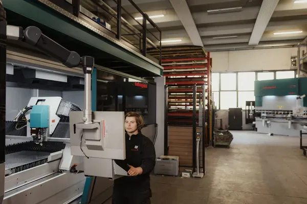 Eine Person bedient eine große Metallbearbeitungsmaschine in einer Werkshalle; im Hintergrund Regale, weitere Maschinen und helles Licht.