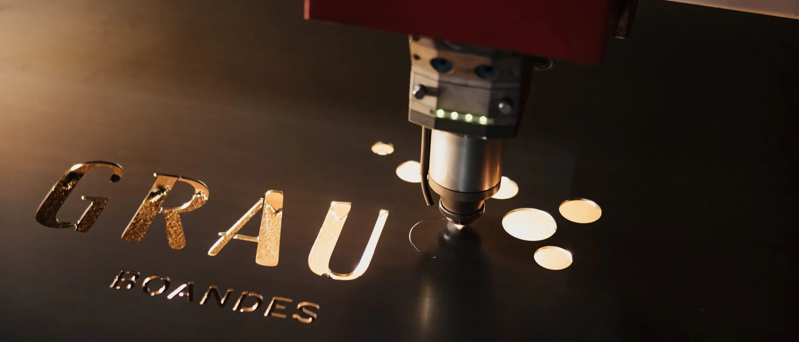 Lasermaschine schneidet Buchstaben 'GRAU' in dunkles Metallblech, neben runden Kreisen; roter Kopf, Südtiroler Metallbaufirma.
