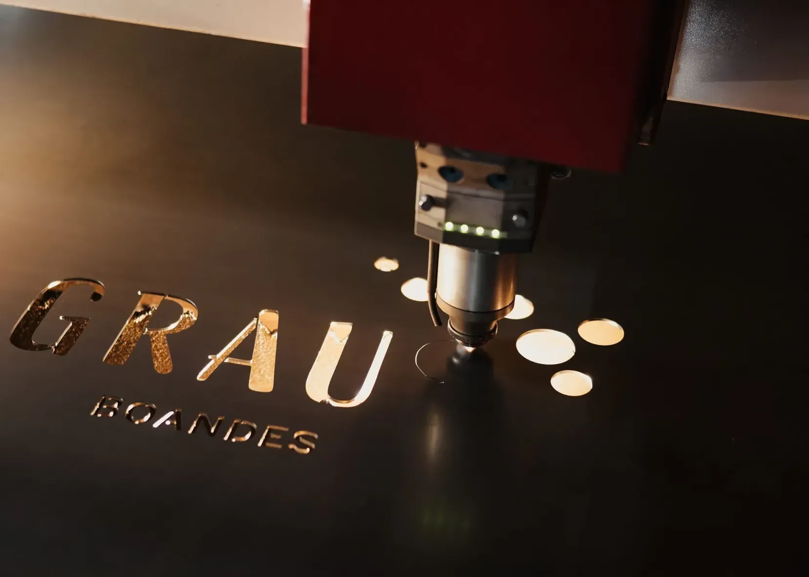 Lasermaschine schneidet Buchstaben 'GRAU' in dunkles Metallblech, neben runden Kreisen; roter Kopf, Südtiroler Metallbaufirma.