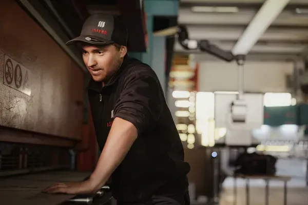 Person in schwarzer Arbeitskleidung und Mütze lehnt an einer Werkbank in Metallwerkstatt in Südtirol und blickt konzentriert auf eine Maschine.