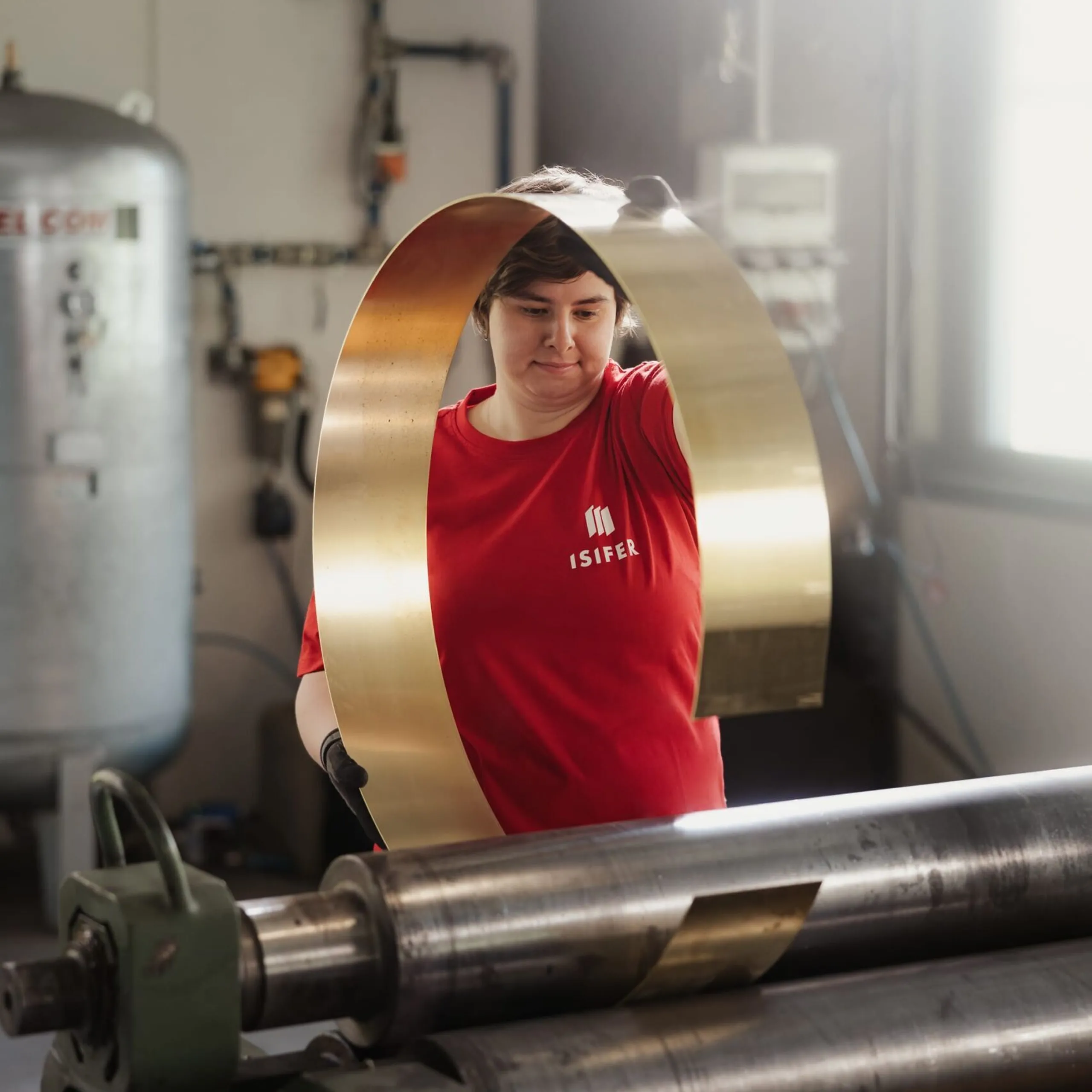 Person in rotem ISIFEK-Shirt in einer südtiroler Metallwerkstatt, hält ein goldfarbenes Metallband um sich, während Walzenmaschinen im Hintergrund laufen.