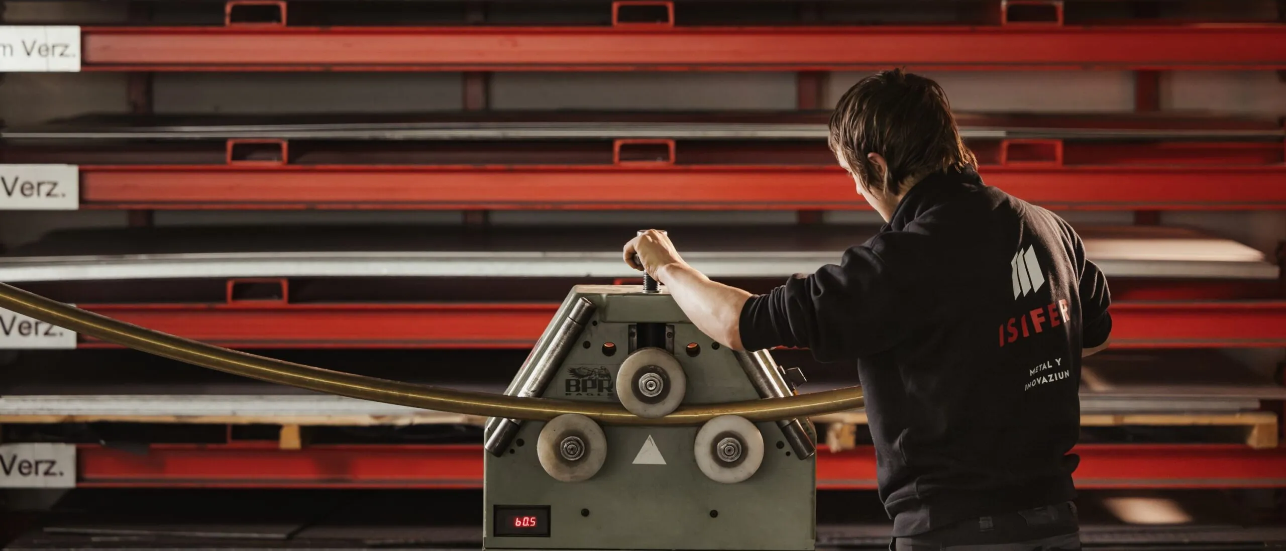 Person bedient eine Blechbearbeitungsmaschine in einer Werkstatt; im Hintergrund rote Metallregale mit gestapelten Blechen.