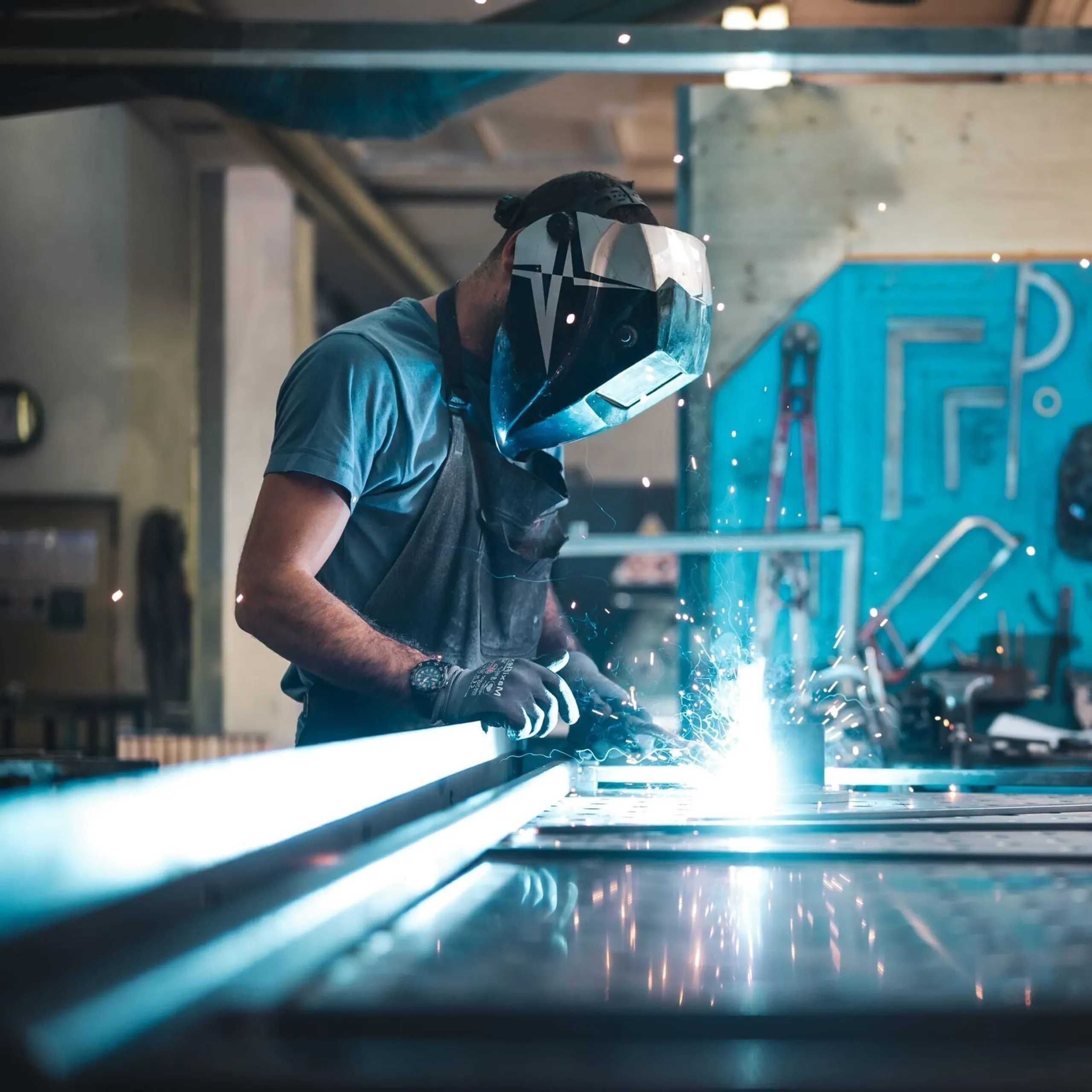 Person mit Schutzhelm und Handschuhen schweißt eine Metallplatte; Funken sprühen, Hintergrund mit Werkzeugen an einer blauen Werkstattwand.