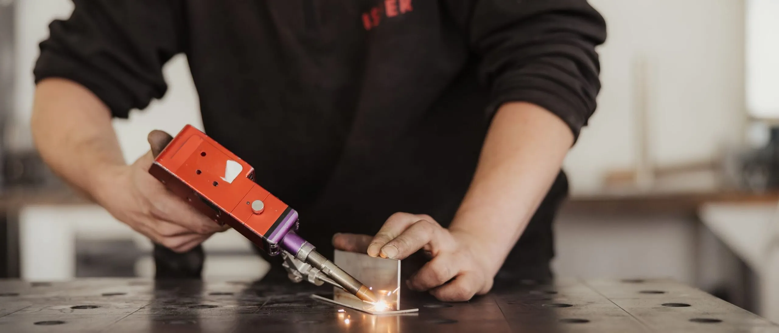 Person in schwarzer Arbeitskleidung schweißt Metall auf einer gelochten Werkbank; Funken sprühen in einer Südtiroler Metallbau-Werkstatt.