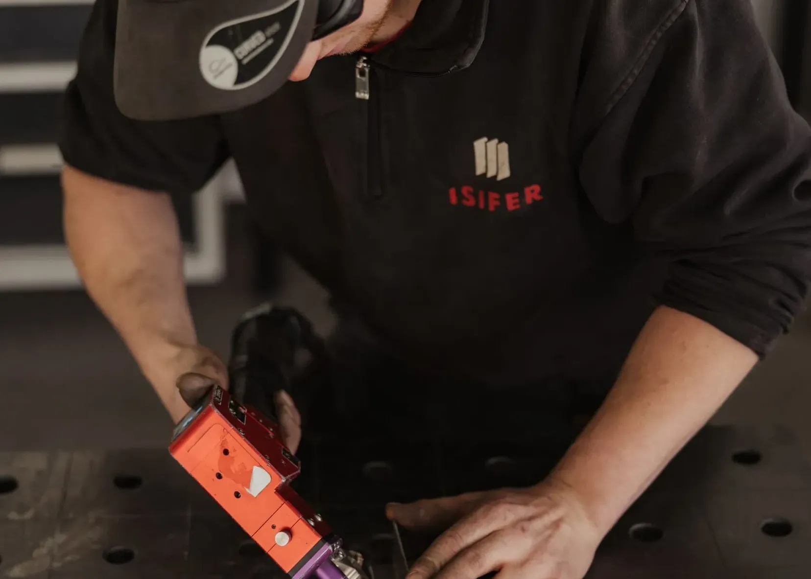 Person aus einem Metallbauunternehmen in Südtirol schweißt ein Blechteil; Funken fliegen, dunkle Kleidung, Kappe und Pullover mit ISIFER-Logo vor einer Werkbank.