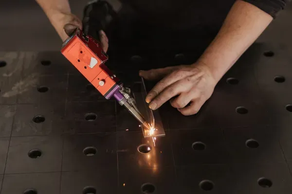 Person aus einem Metallbauunternehmen in Südtirol schweißt ein Blechteil; Funken fliegen, dunkle Kleidung, Kappe und Pullover mit ISIFER-Logo vor einer Werkbank.