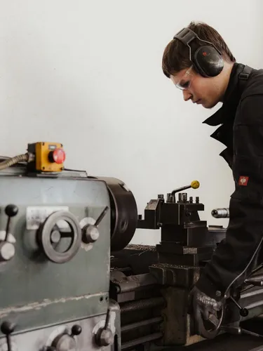 Eine Person in dunkler Arbeitskleidung bedient eine Metall-Drehbank in einer Werkstatt, trägt Gehörschutz, Schutzbrille und Handschuhe.