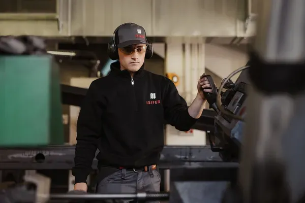 Person im schwarzen Pullover mit Isifer-Logo bedient eine Metallmaschine, trägt Schutzbrille und Gehörschutz in einer Fertigungshalle.