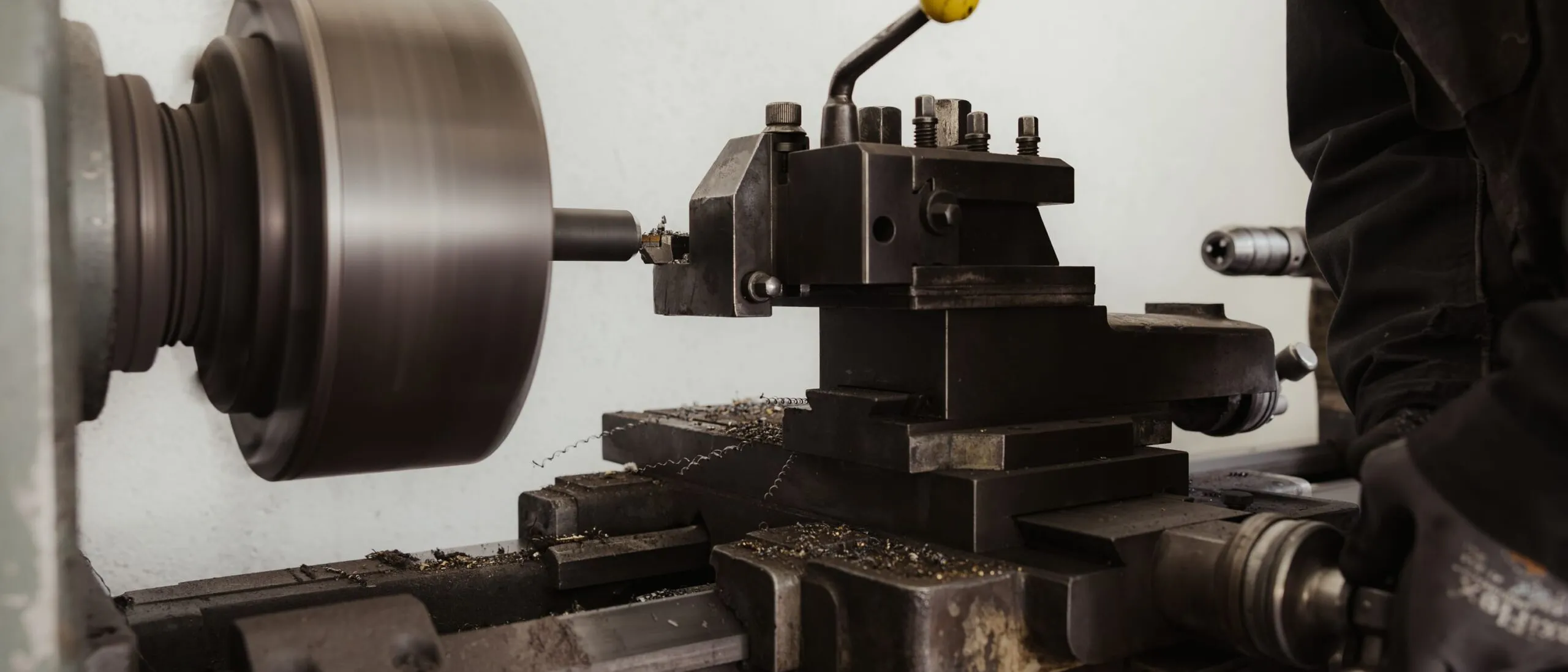 Metall-Drehmaschine in einer Südtiroler Werkstatt; rotiertes Werkstück wird bearbeitet, Späne liegen auf dem Maschinentisch, rechts eine Person mit Handschuh greift zu.