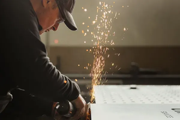 Person in einer Metallwerkstatt schleift Metallplatte mit Winkelschleifer; Funken sprühen, Schutzbrille und Kappe sichtbar.