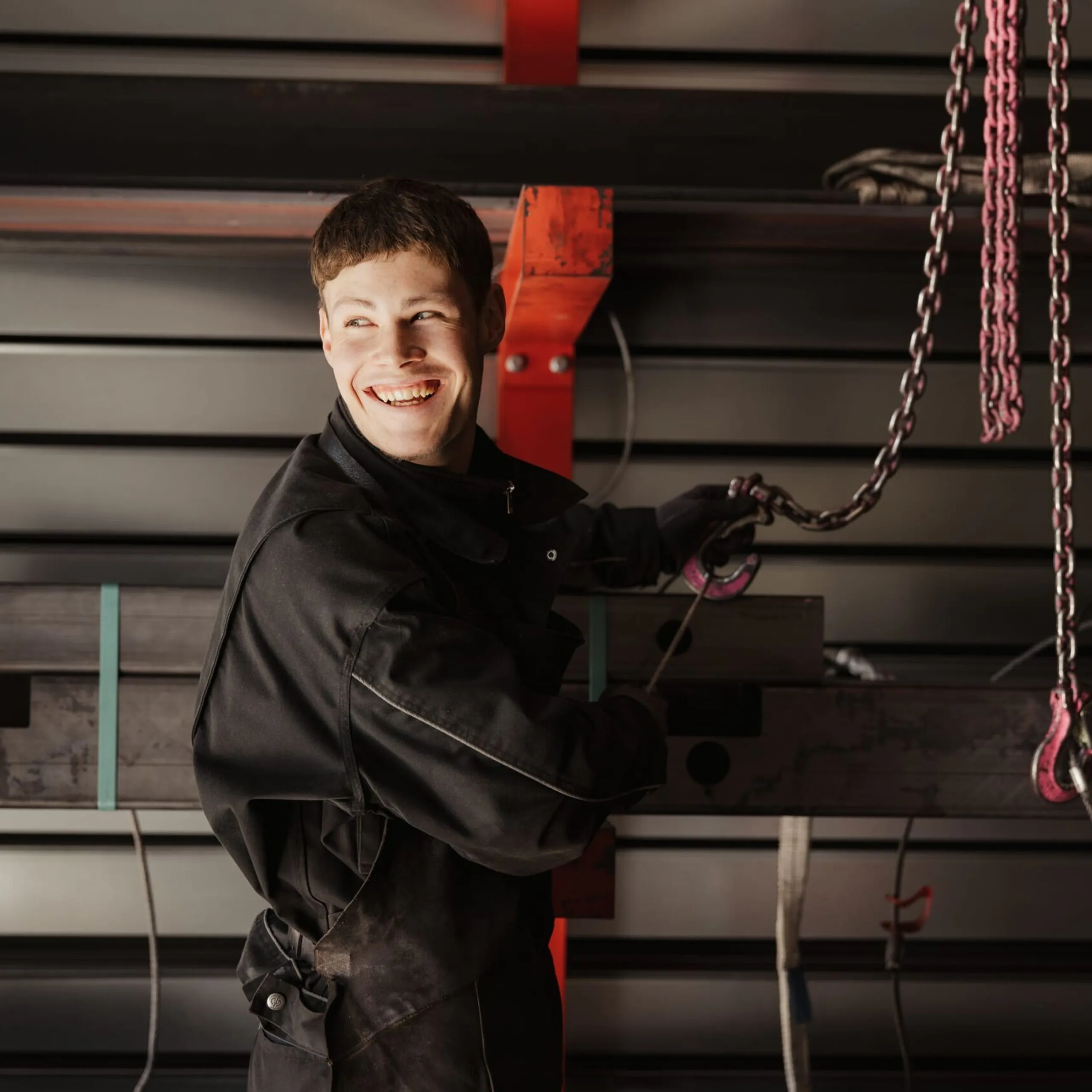 Person in schwarzer Arbeitskleidung in einer Metallwerkstatt, lächelnd, hält eine Kette an schweren Stahlträgern; Hintergrund mit Metallplatten und Hebezeugen.