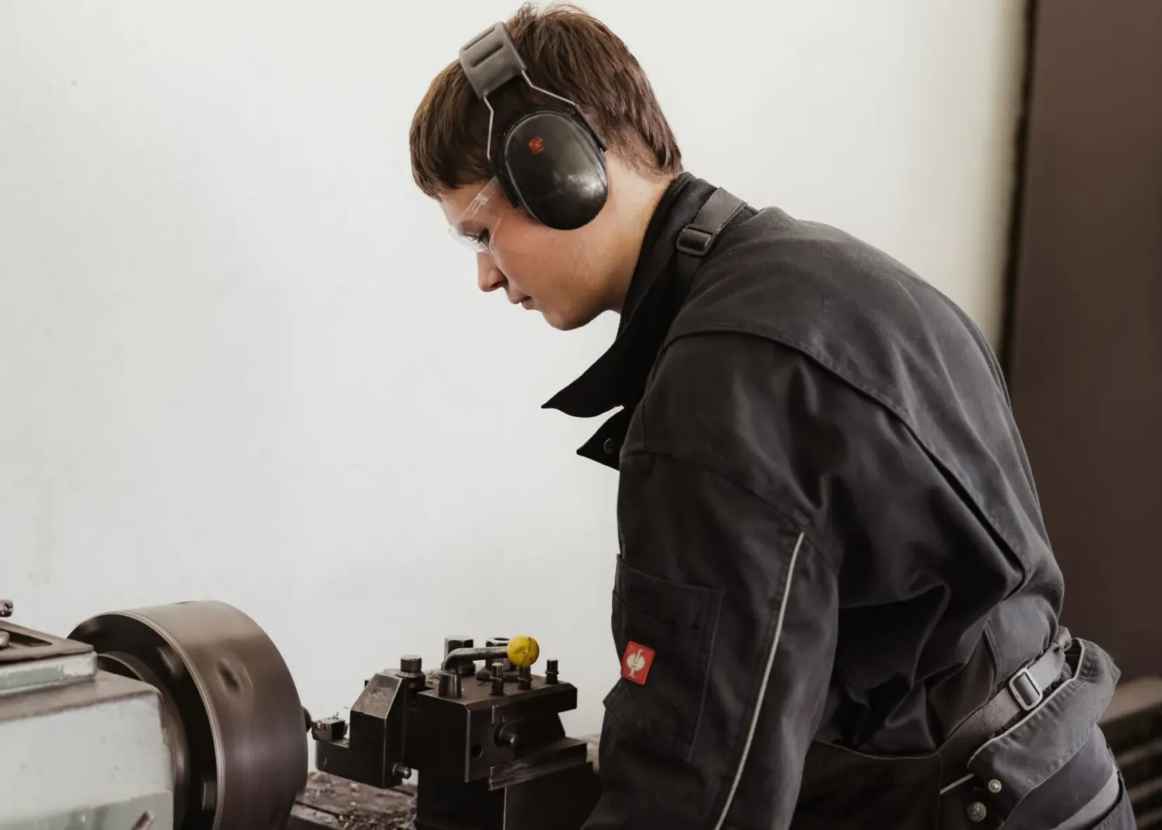 Person in schwarzer Arbeitskleidung mit Gehörschutz und Schutzbrille bedient eine Metall-Drehmaschine; Späne liegen auf dem Maschinentisch.