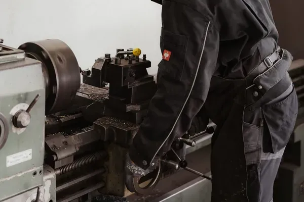 Person in schwarzer Arbeitskleidung mit Gehörschutz und Schutzbrille bedient eine Metall-Drehmaschine; Späne liegen auf dem Maschinentisch.