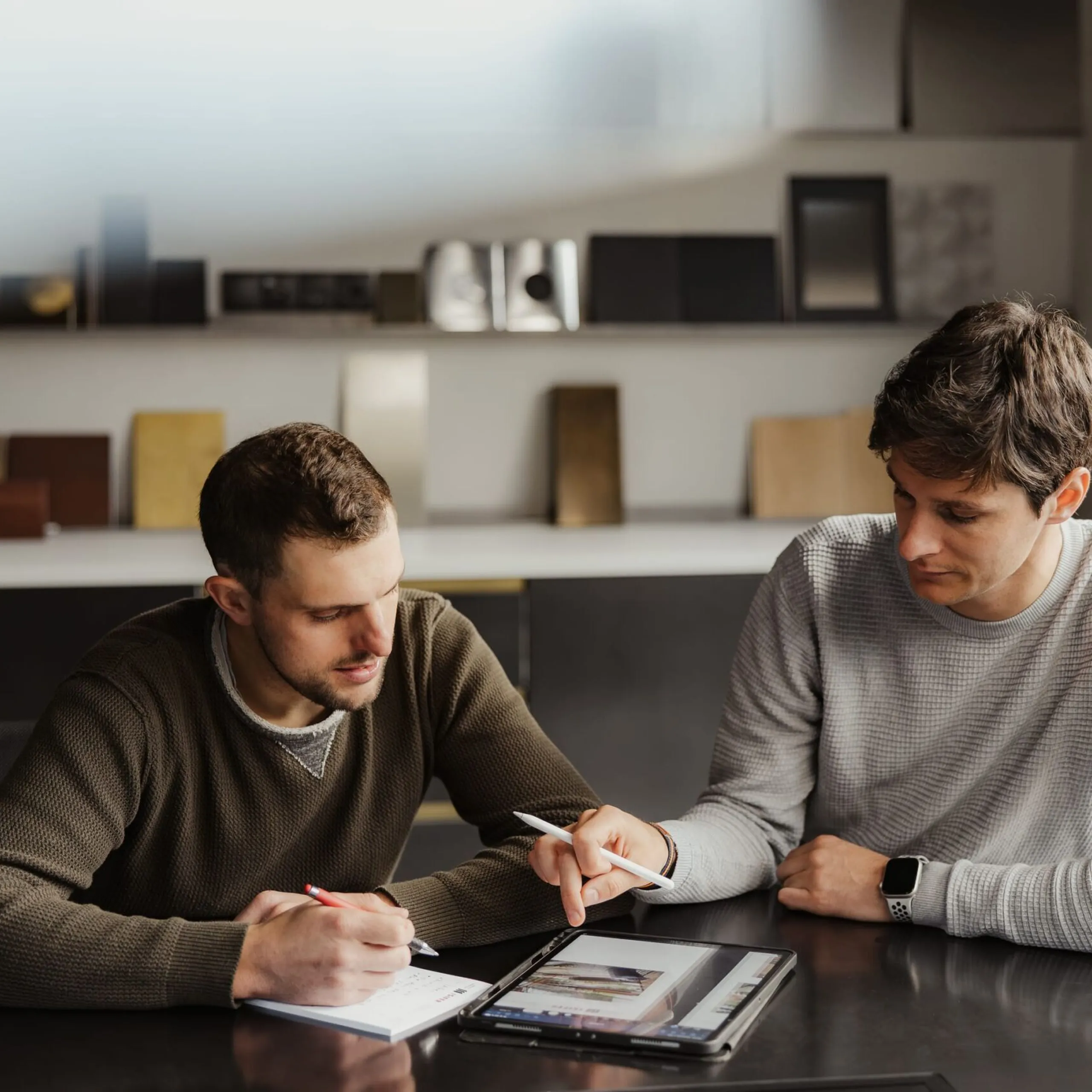Zwei Mitarbeitende sitzen an einem dunklen Tisch, notieren Dinge und arbeiten am Tablet, während Metallproben im Hintergrund ausgestellt sind.