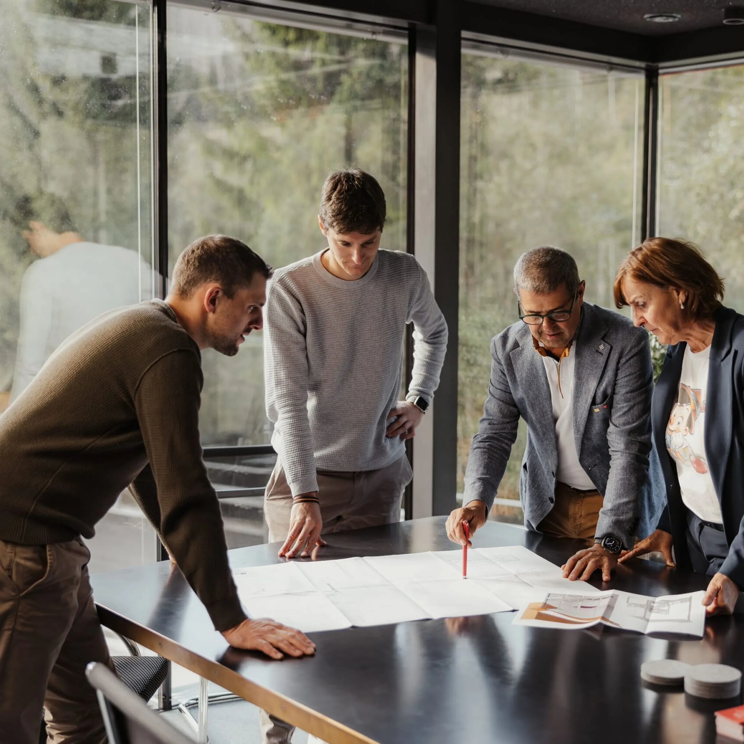 Vier Personen diskutieren Baupläne auf einem Konferenztisch in einem Glas-Konferenzraum; draußen sind grüne Bäume zu sehen.