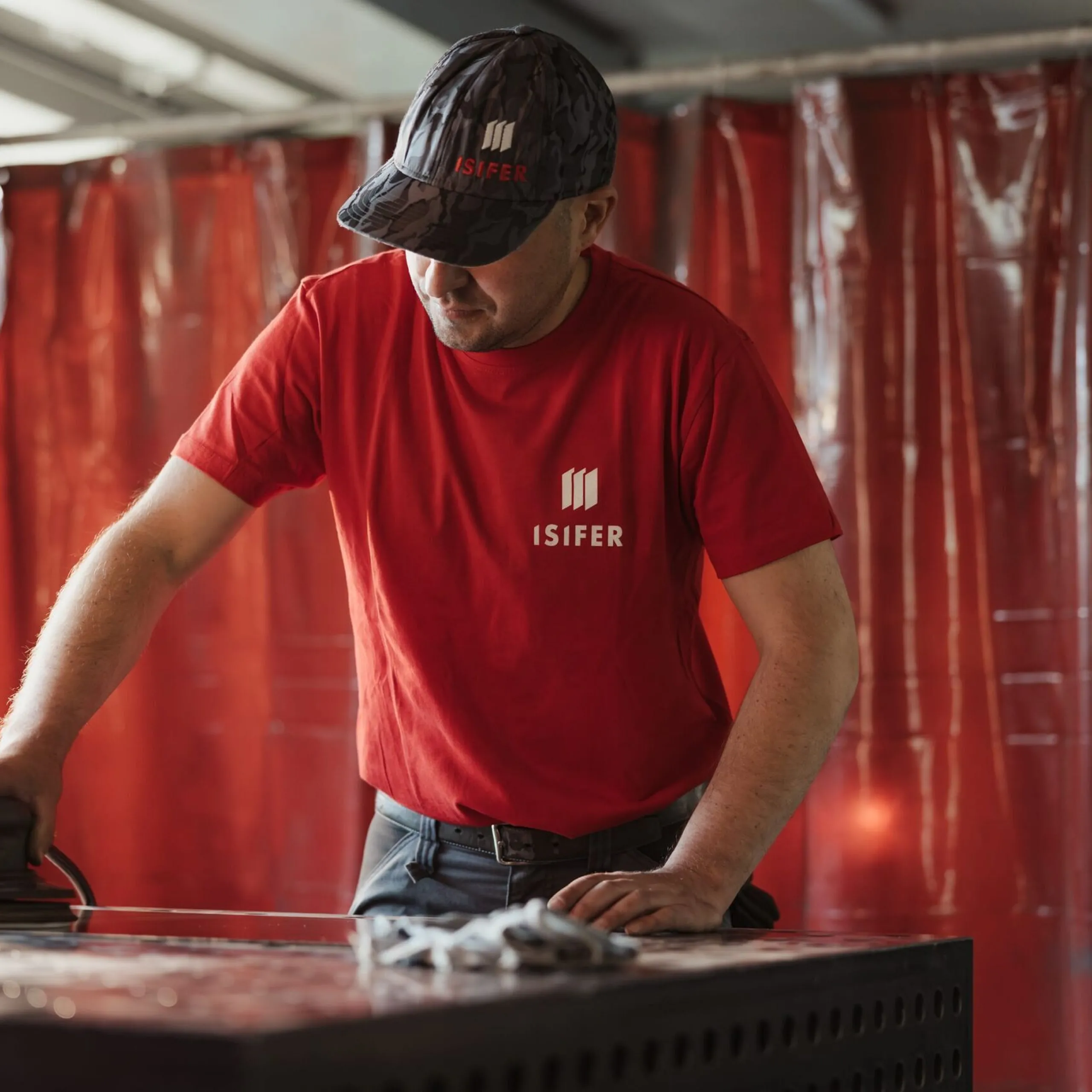 Person in rotem ISIFER-T-Shirt bearbeitet eine Metallfläche mit einem Schleifgerät in einer Werkstatt; rote Trennvorhänge im Hintergrund.
