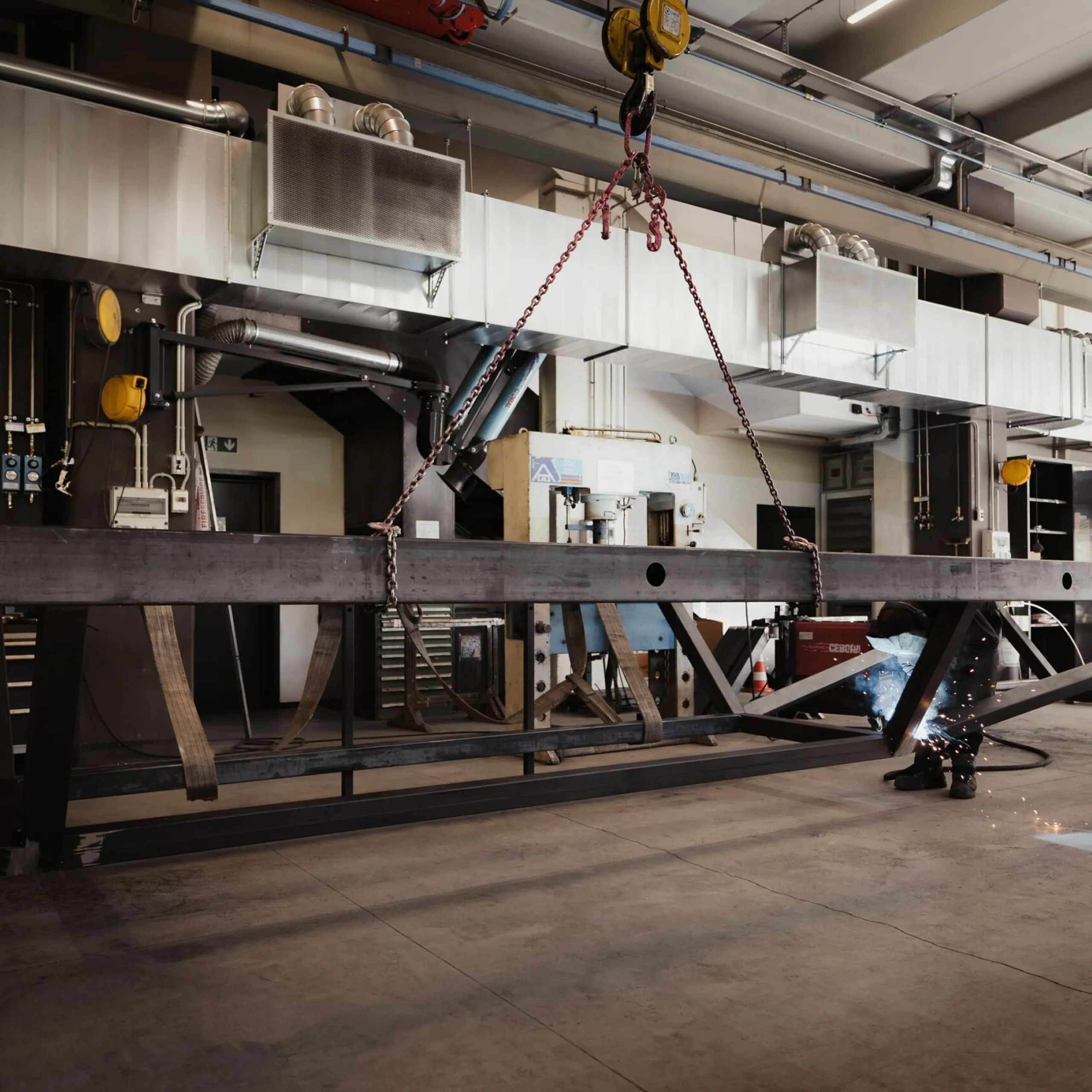 Große Metallbau-Werkhalle in Südtirol; eine Person schweißt an einer langen Stahlträgerkonstruktion, Funken fliegen; Hintergrund: Maschinen.