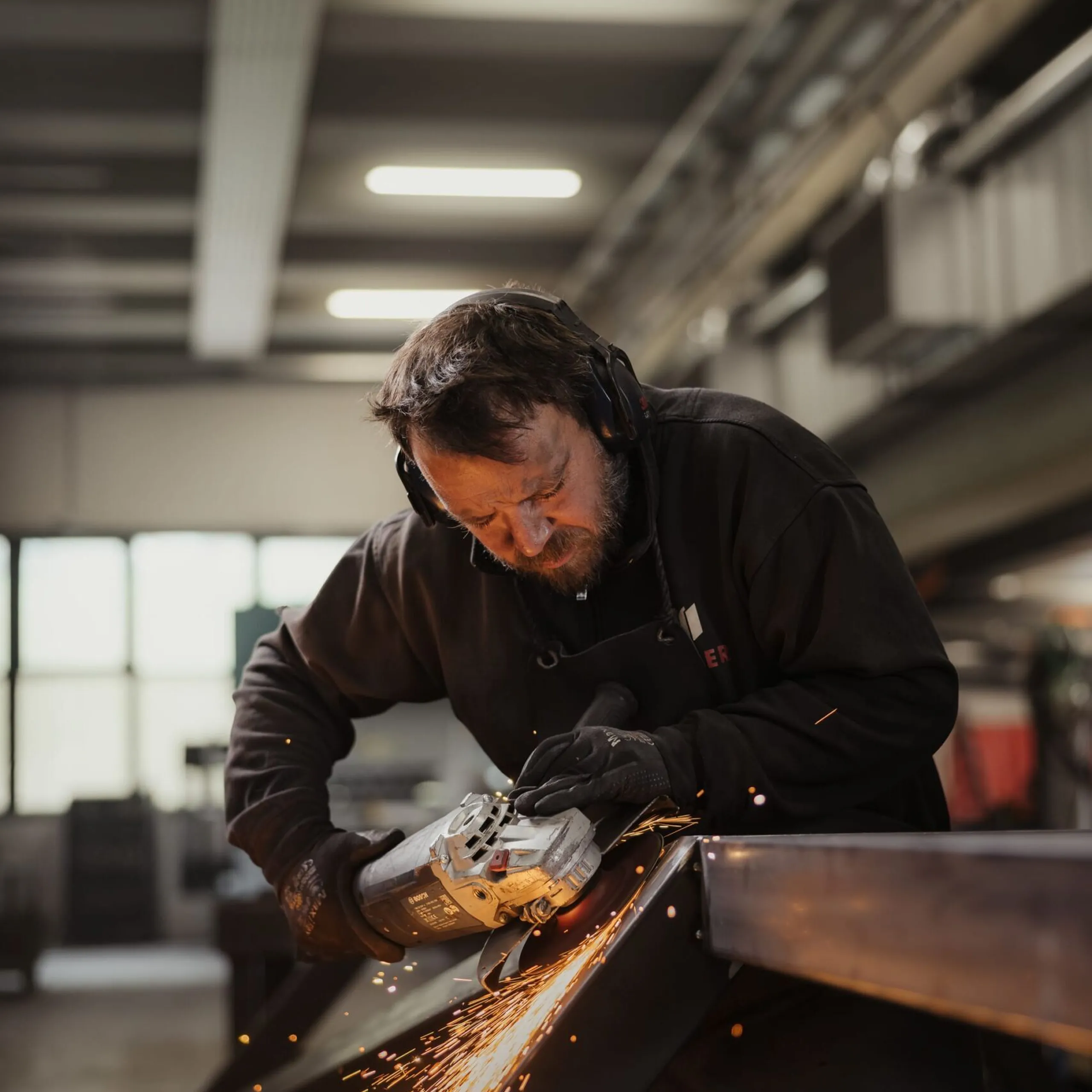 Person mit Gehörschutz und Handschuhen bearbeitet einen Metallträger mit einem Winkelschleifer; Funken fliegen in einer Industriehalle.