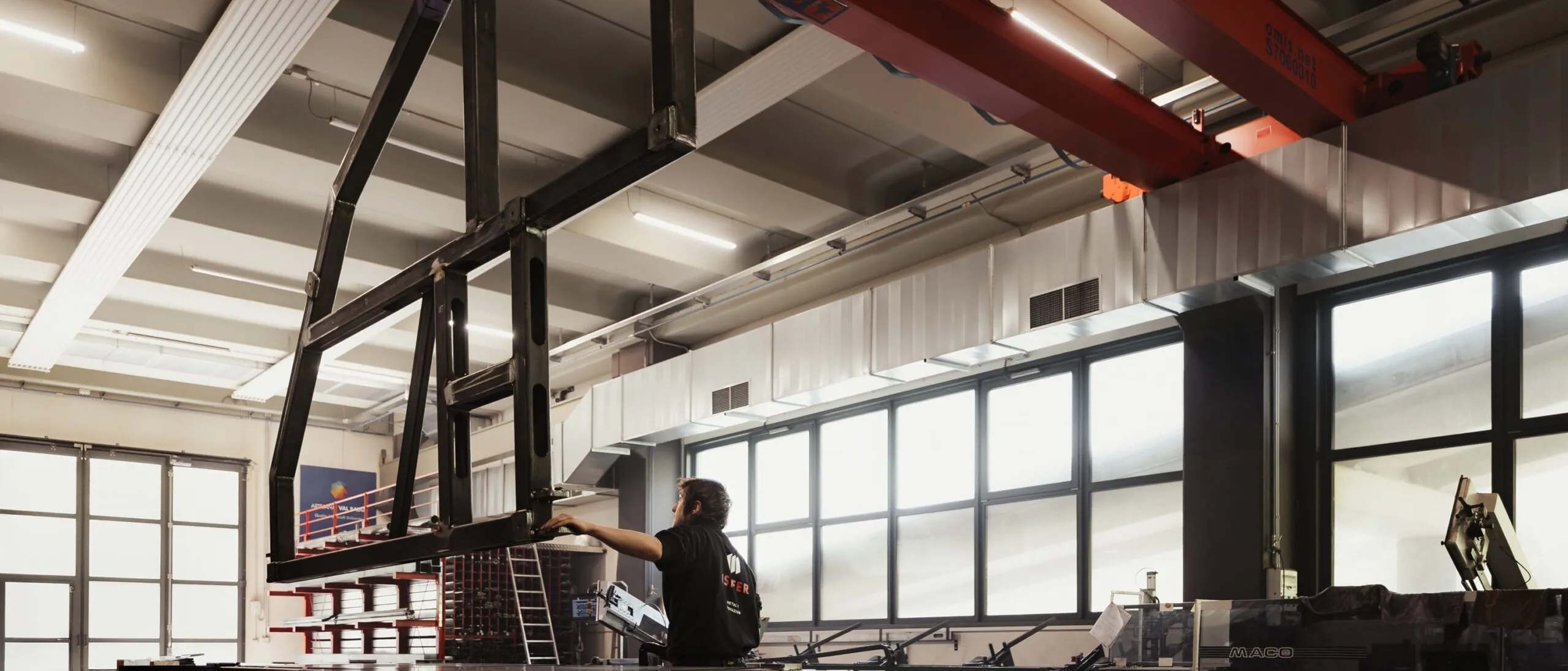 Metallbauwerk in einer Südtiroler Industriehalle: schwarzes Rahmengestell hängt am roten Hallenkran, eine Person montiert es vor großen Fenstern.