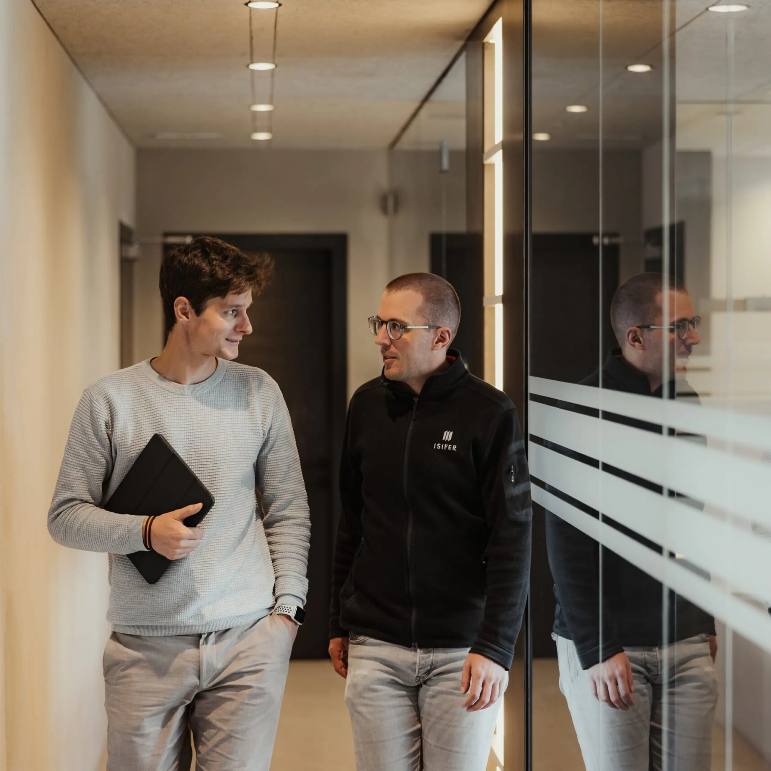 Zwei Personen in einem modernen Büroflur mit Glaswänden; eine hält einen schwarzen Ordner, die andere trägt eine Jacke mit Logo, beide im Gespräch.