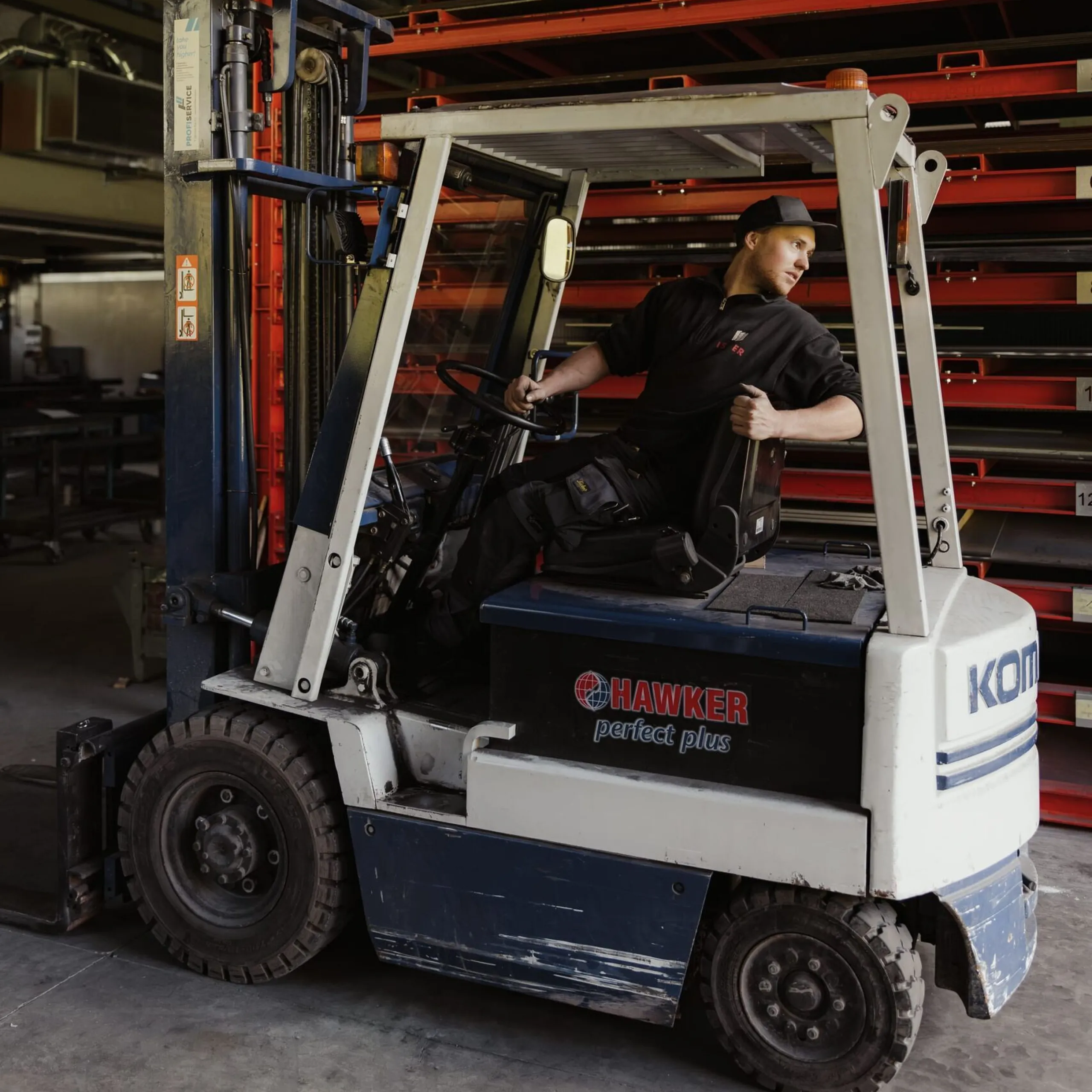 Eine Person fährt einen Hawker-Gabelstapler in einer Werkstatt einer Metallbaufirma in Südtirol, umgeben von hohen roten Metallregalen.