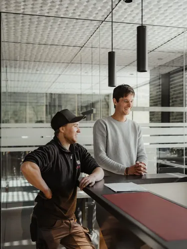 Drei Personen an einer Rezeption in einer Metallbau-Firma in Südtirol; Glaswände, dunkle Leuchten, Notiz auf dem Tresen, freundliches Gespräch.