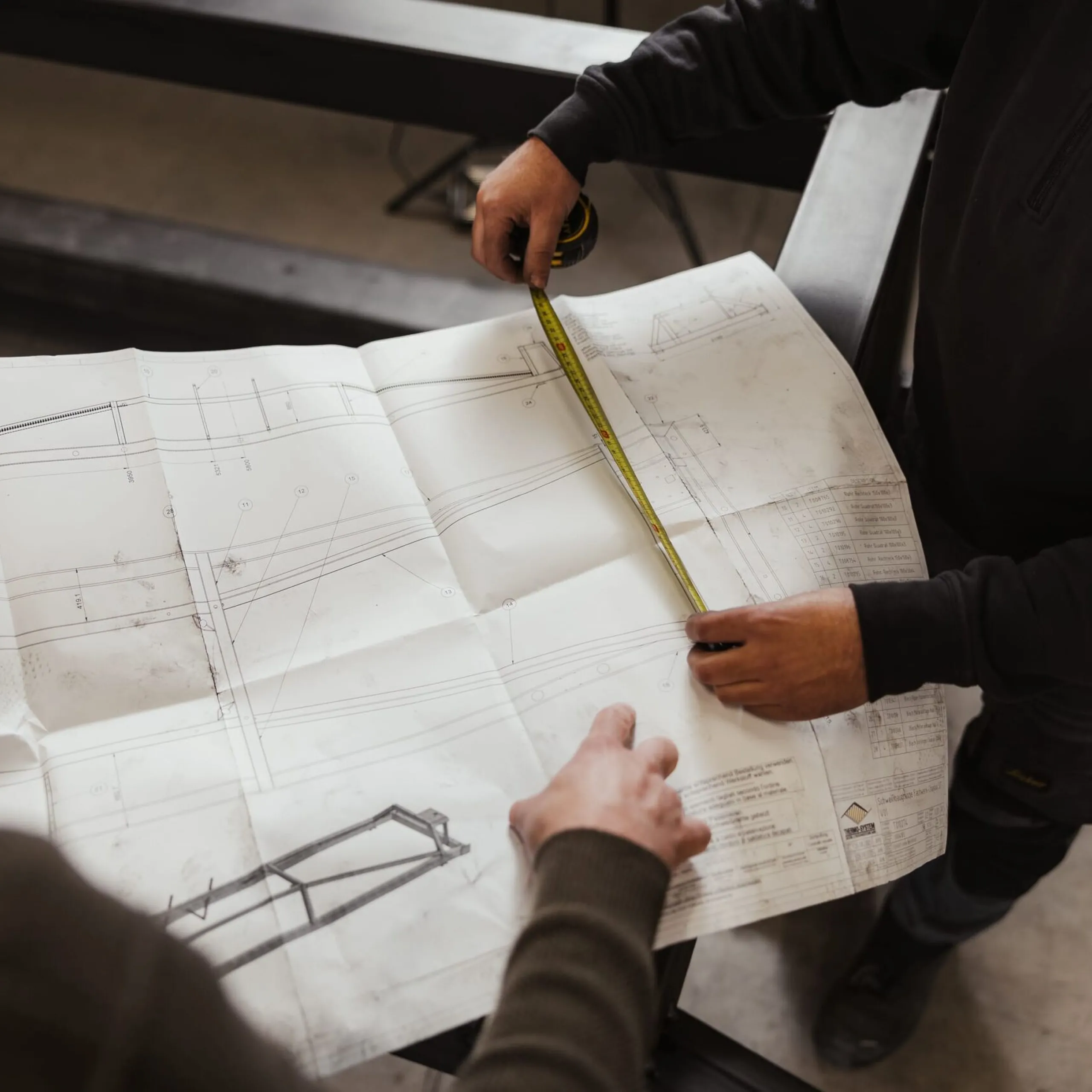 Zwei Mitarbeitende eines Südtiroler Metallbauunternehmens prüfen eine große Bauzeichnung, während eine Person mit Maßband darüber misst.