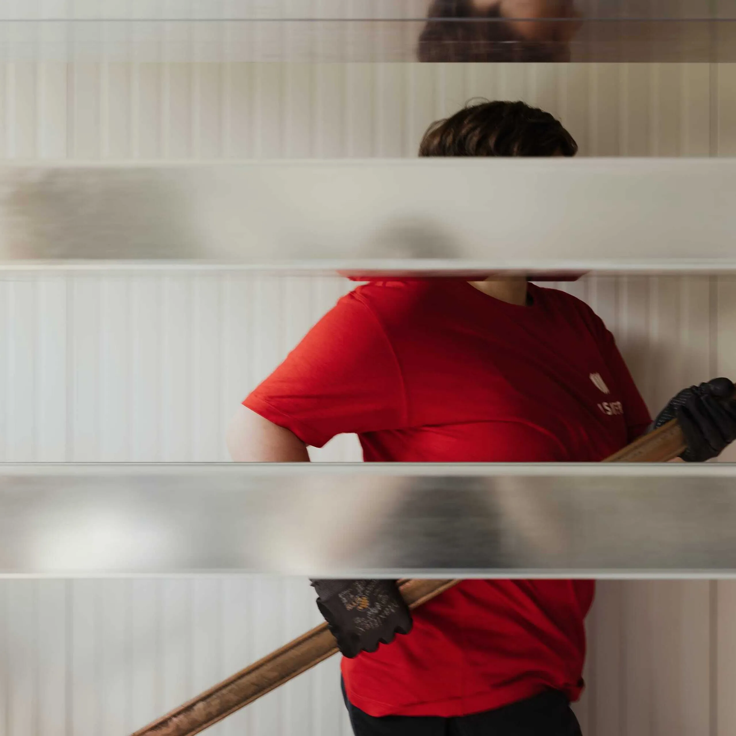 Person in rotem Shirt und Handschuhen hält ein langes Metallrohr vor einer hellen Paneelwand; horizontale Metallbalken schneiden die Sicht.