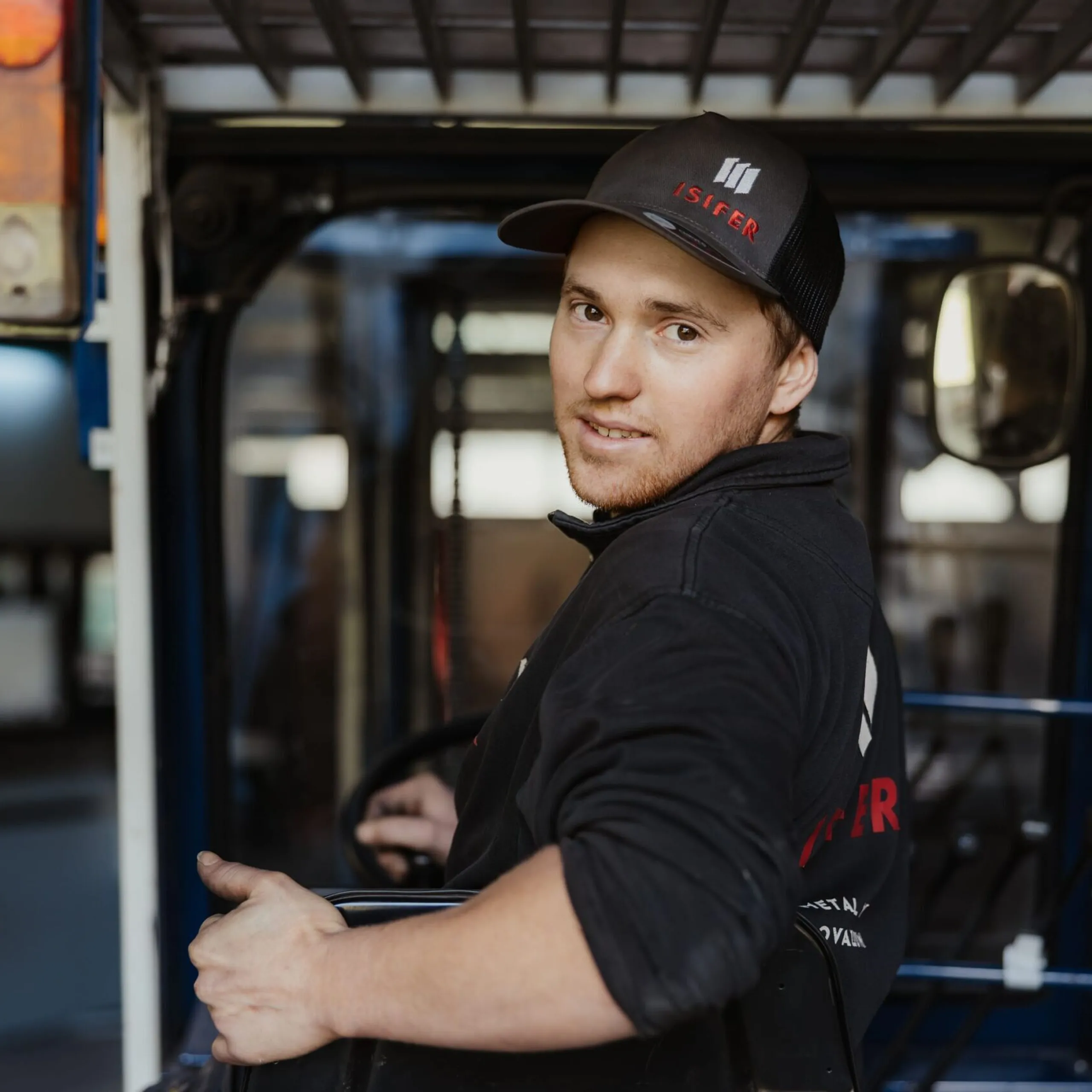 Person in Arbeitskleidung sitzt im Führerstand eines Fahrzeugs, blickt in die Kamera; schwarze Jacke, Cap, Werkstatt im Hintergrund.