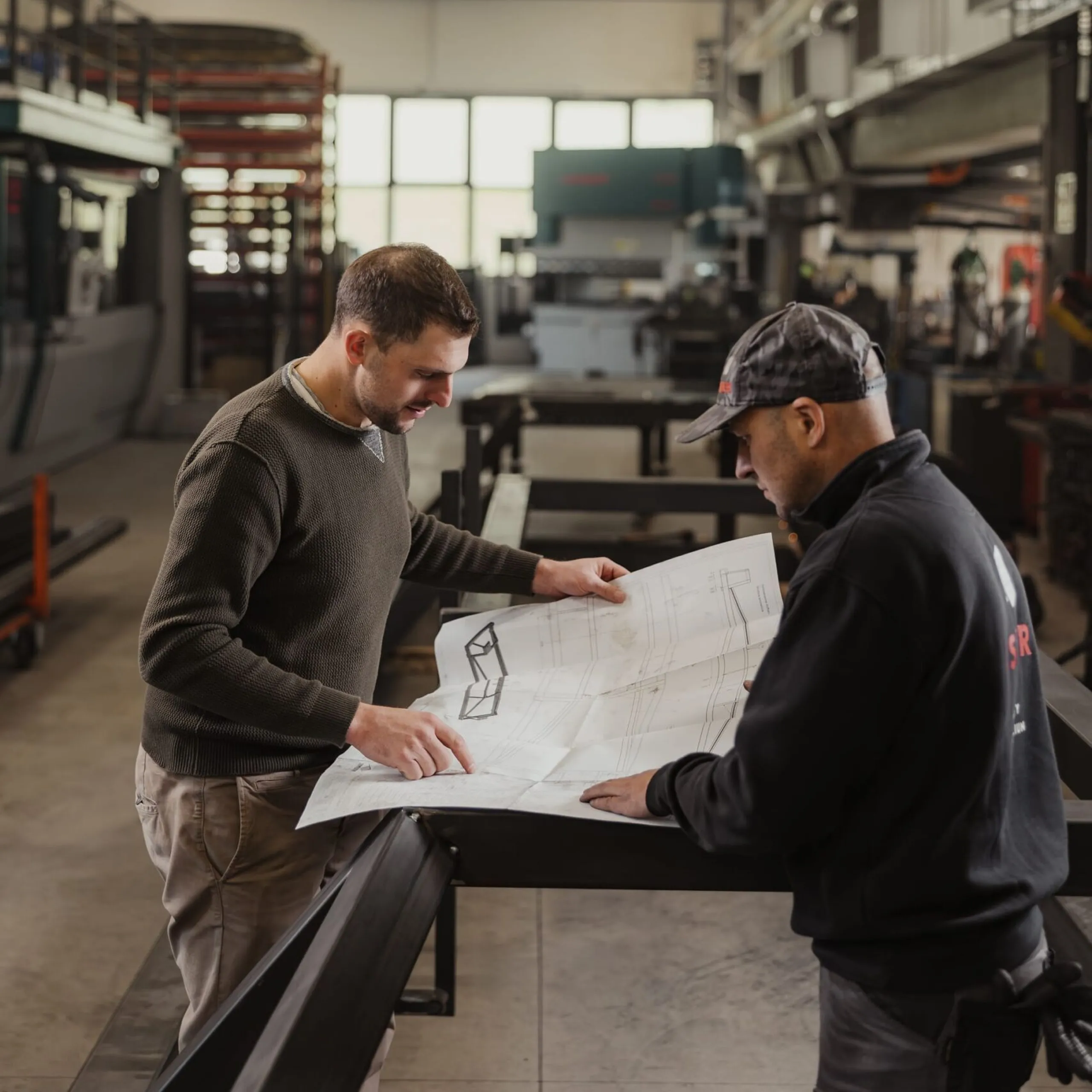 Zwei Mitarbeitende eines Südtiroler Metallbaubetriebs prüfen in einer hellen Werkstatthalle einen Bauplan auf einem Tisch, umgeben von Maschinen.