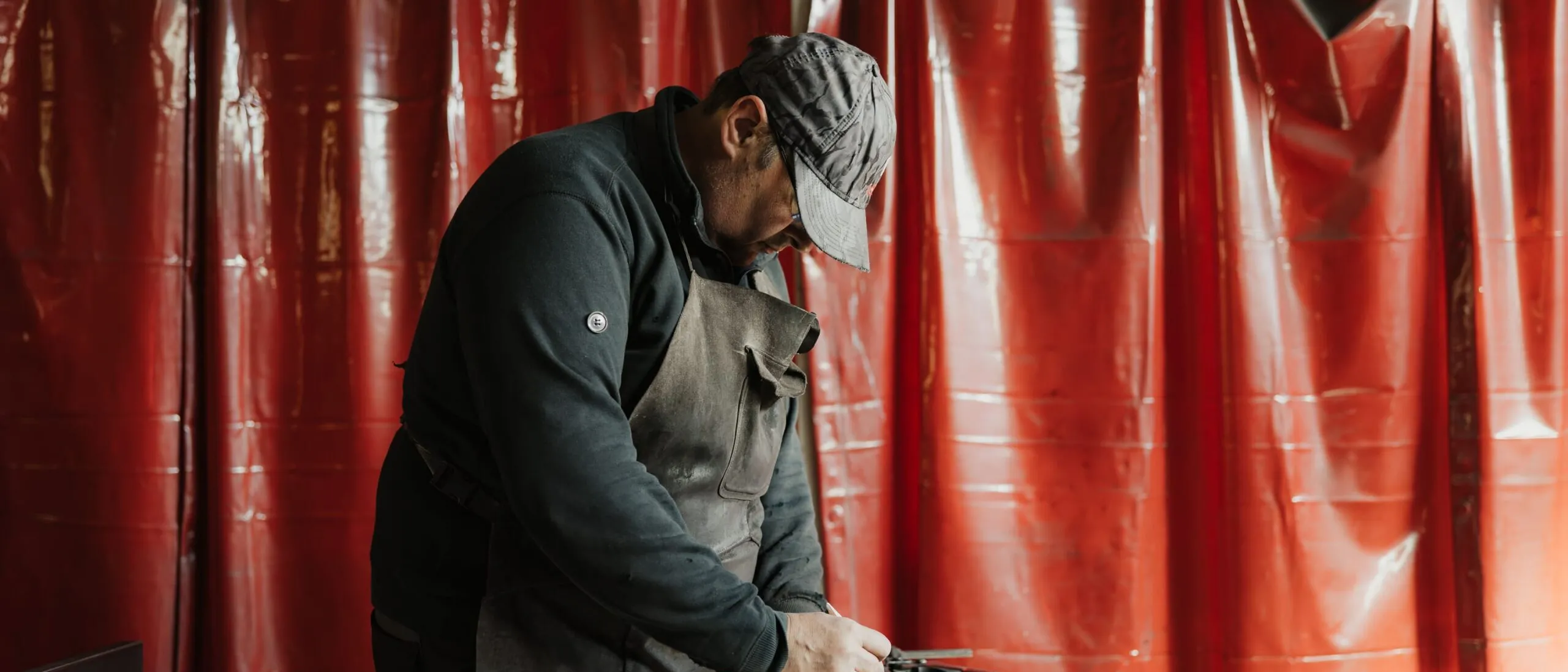 Una persona con cappello e grembiule lavora su una barra di metallo sul banco da lavoro in un'officina, sfondo tende rosse.