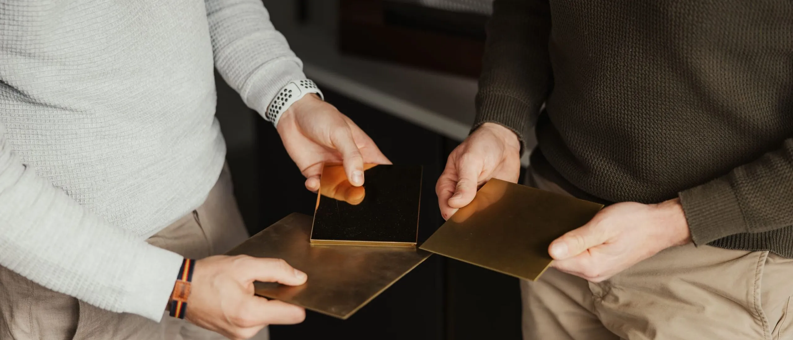 Zwei Personen halten Messingplatten und vergleichen Metallproben in einer Werkstatt.