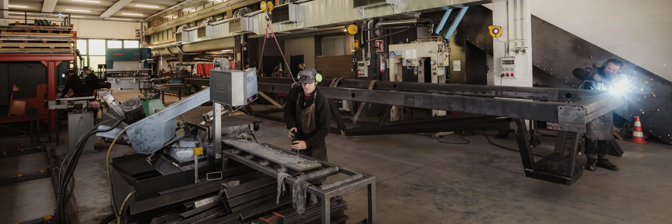 Metallbau-Werkstatt in Südtirol: Mitarbeitende bearbeiten große Stahlträger, sichtbar sind Schweißarbeiten, Maschinen und ein roter Hallenkran.