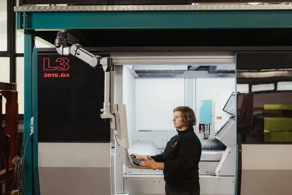 Person in dunkler Kleidung bedient eine CNC-Fertigungseinheit in einem Südtiroler Metallbauunternehmen; Roboterarm neben offenem Maschinenraum, Steuerpult, Beschriftung L3 3015.G4.