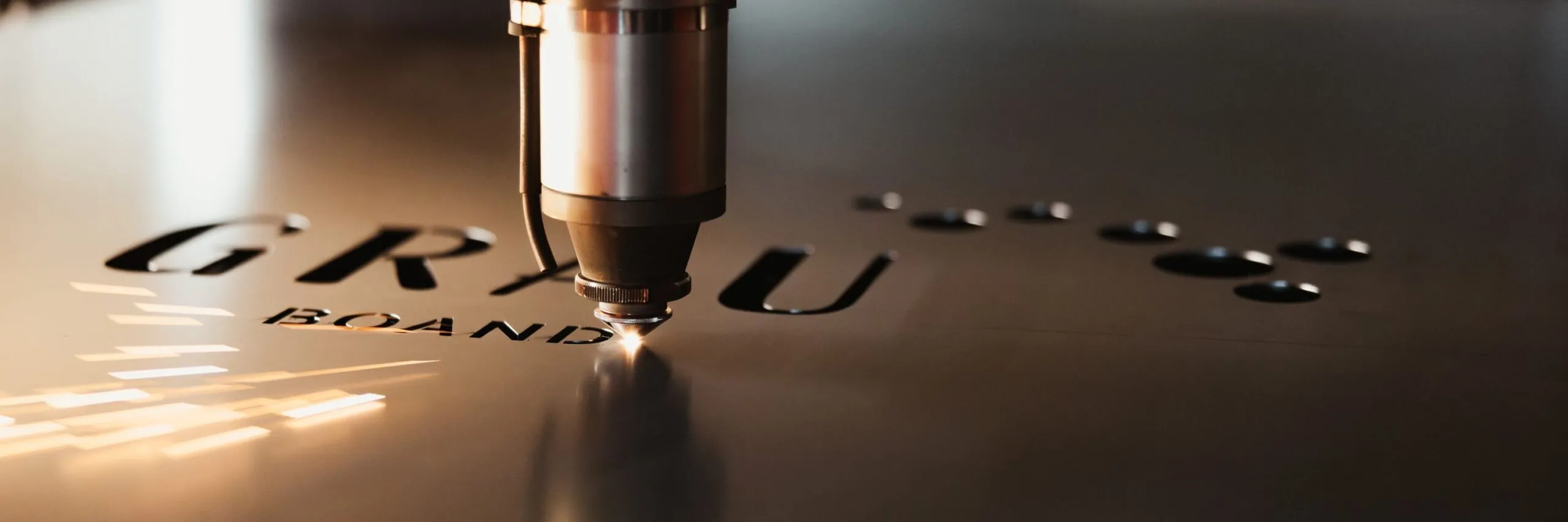Nahaufnahme eines Laser-Schneidkopfs beim Gravieren einer Metallplatte; markante Buchstaben des Firmenlogos, Funkenflug und spiegelnde Metalloberfläche.