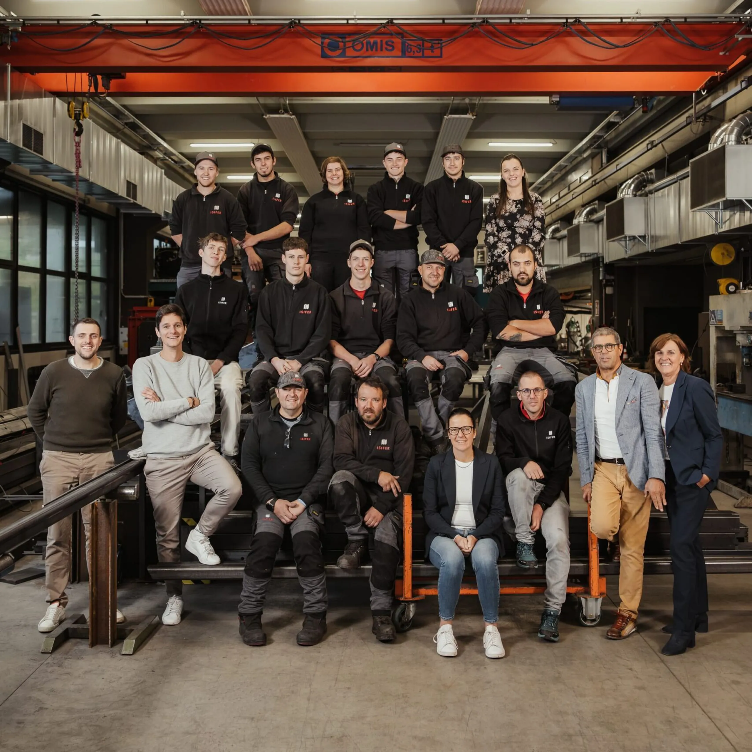 Gruppenfoto von Mitarbeitenden eines Metallbau-Unternehmens in Südtirol, versammelt in einer Werkstatt auf Stahlkonstruktionen, vor einem orangefarbenen Hallenkran.