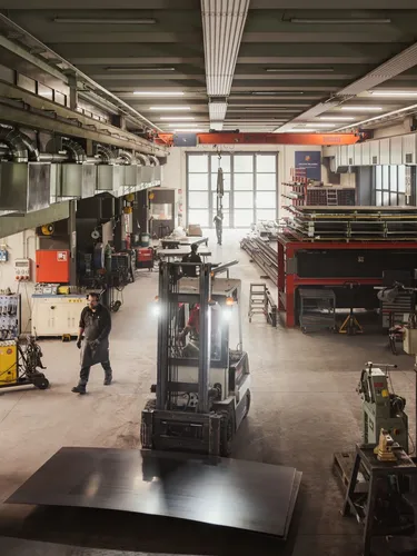 Große Metallwerkstatt in Südtirol, Gabelstapler hebt eine Stahlplatte; rechts rote Regale mit Metallbauteilen, links Maschinen und Arbeiter bei der Arbeit.