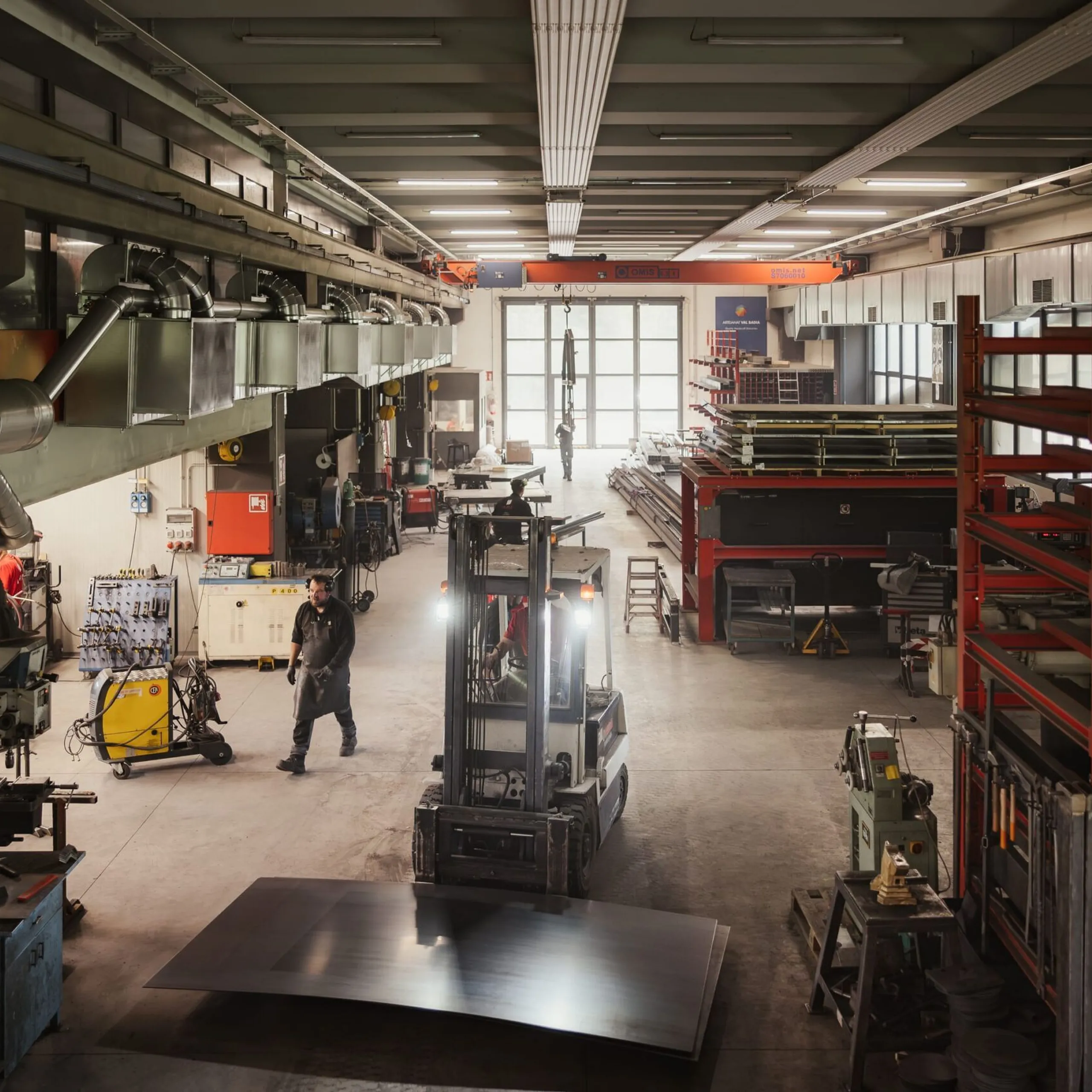 Große Metallwerkstatt in Südtirol, Gabelstapler hebt eine Stahlplatte; rechts rote Regale mit Metallbauteilen, links Maschinen und Arbeiter bei der Arbeit.