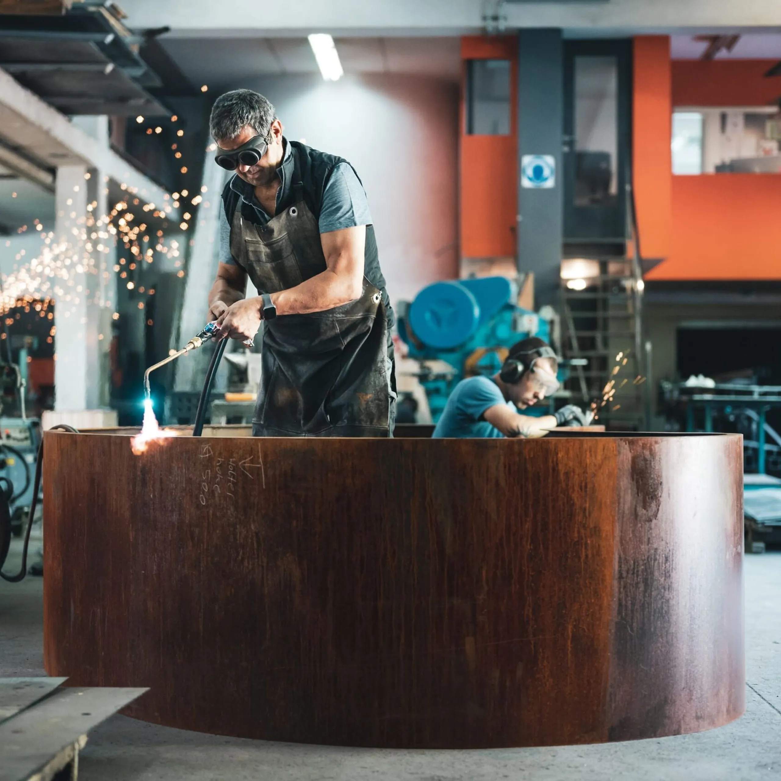 In einer Metallwerkstatt schweißt eine Person an einer großen runden Stahltonne; Funken fliegen, im Hintergrund arbeitet eine weitere Person.