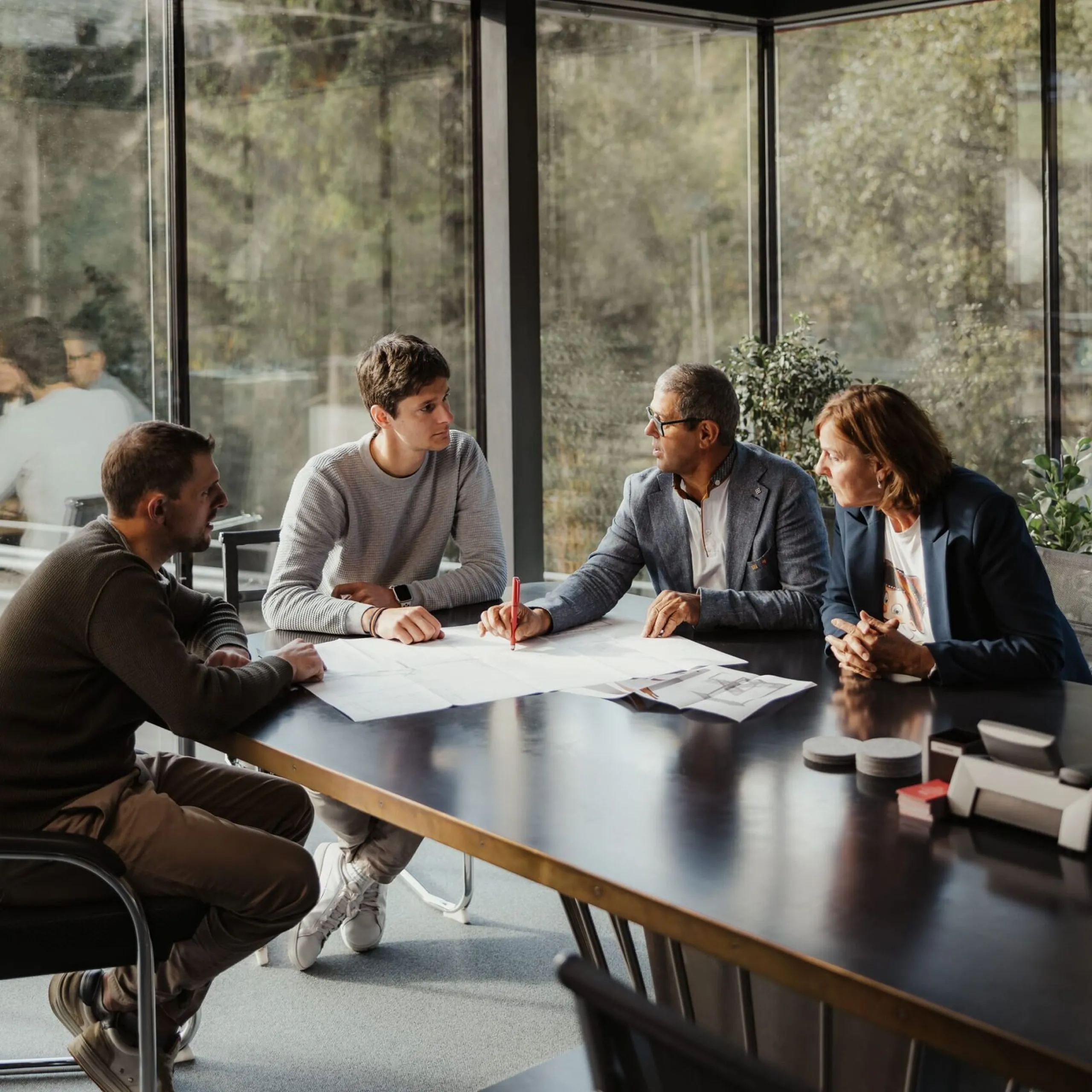 Vier Personen diskutieren Unterlagen an einem Tisch in einem modernen Konferenzraum eines Metallbauunternehmens in Südtirol, große Fenster, reflektierende Decke.