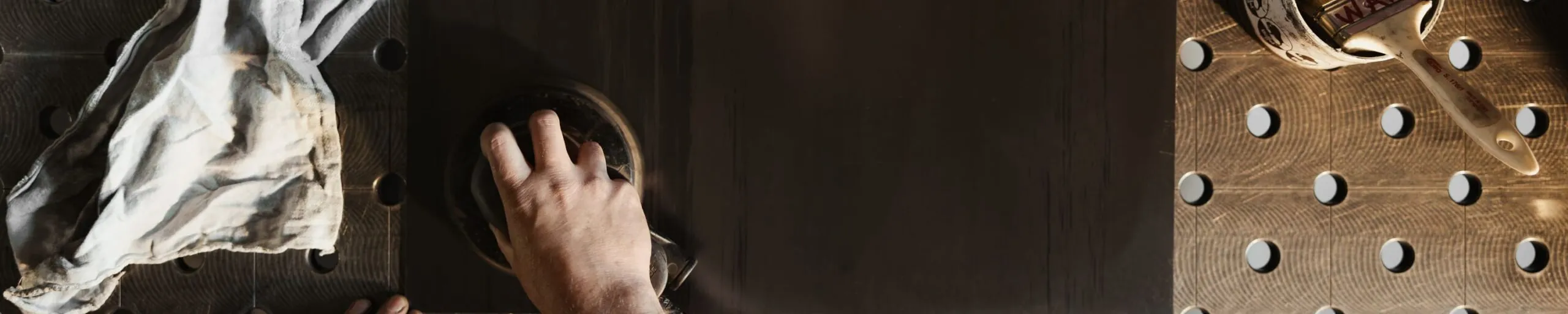 Nahaufnahme: Eine Person poliert einen schwarzen Metallblock auf einer gelochten Metall-Arbeitsfläche; links liegt ein Lappen, rechts ein Becher mit Wachs.