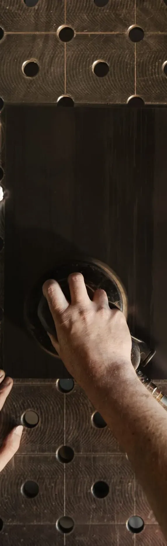 Nahaufnahme: Eine Person poliert einen schwarzen Metallblock auf einer gelochten Metall-Arbeitsfläche; links liegt ein Lappen, rechts ein Becher mit Wachs.