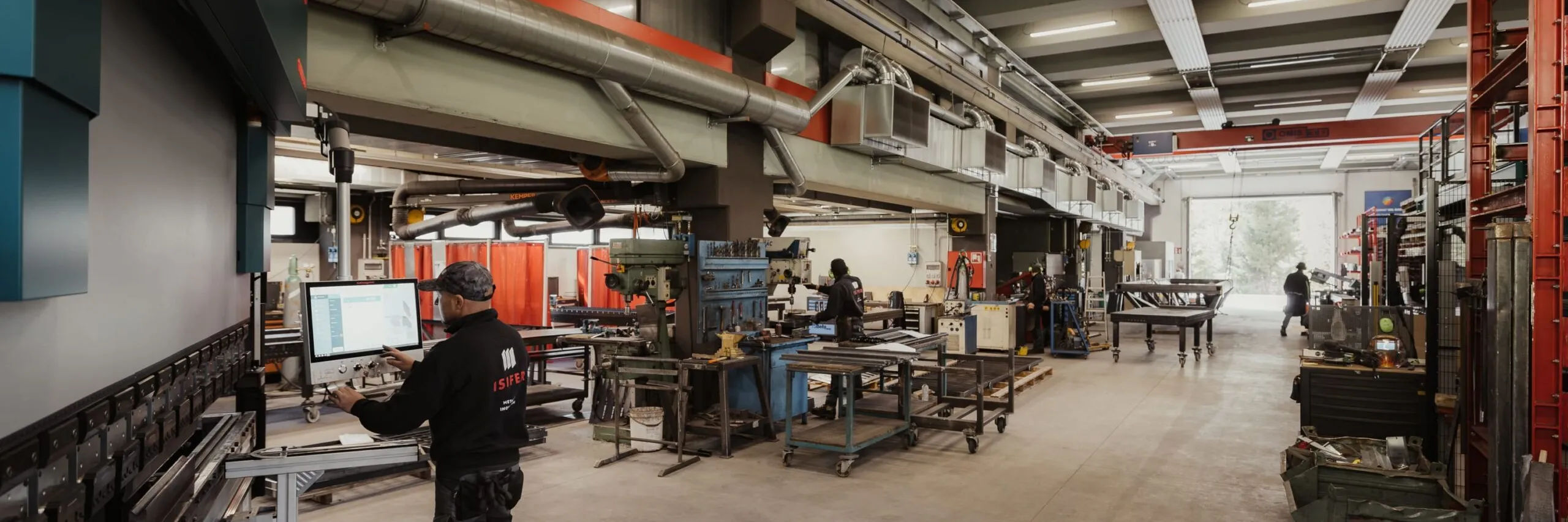 Metallbau-Werkstatt in Südtirol, große Bearbeitungsmaschinen, Werkbänke und Leitungen; eine Person arbeitet am Computer, weitere Mitarbeitende im Hintergrund.