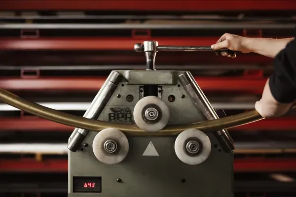 Rohrbiegemaschine in einer Metallwerkstatt; eine Person bedient den Hebel, ein gebogenes Rohr führt durch Rollen, rote Regale im Hintergrund.